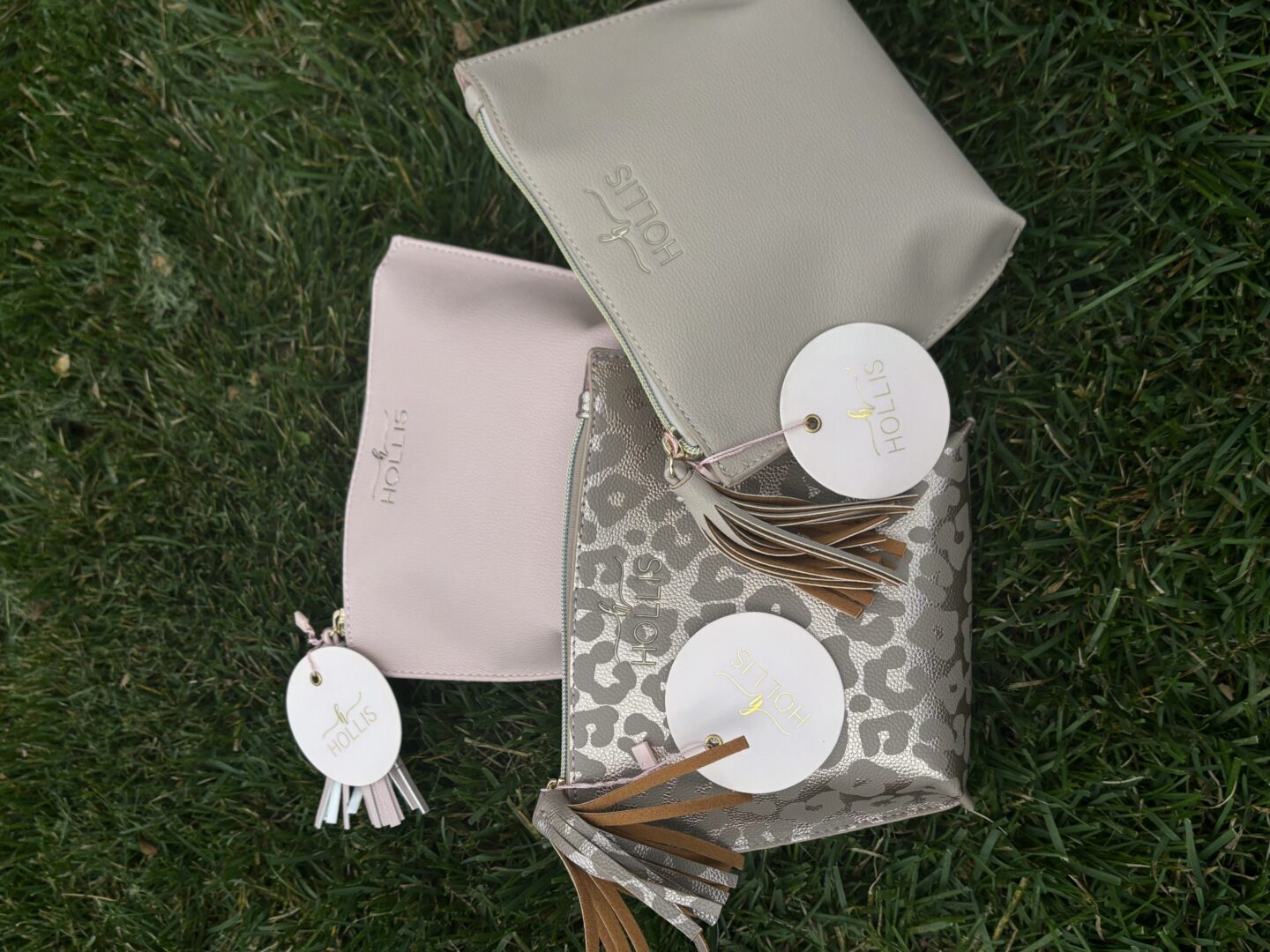 Three Hollis Chic Makeup Travel Bags with tags on them laying on the grass.